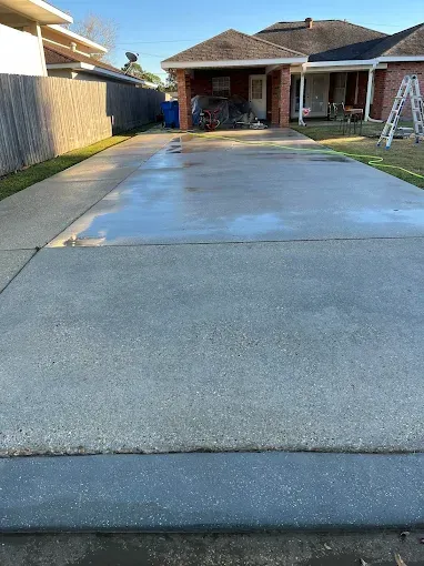 A wet, freshly-cleaned concrete driveway leads to a red brick house. A wooden fence and grass are visible on the left.