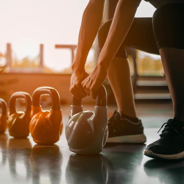 A person in a gym squats to lift a kettlebell, with additional kettlebells lined up on the floor nearby.