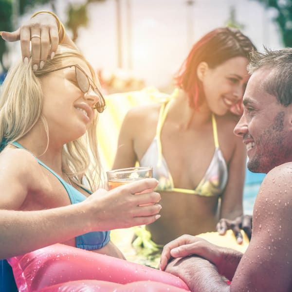 Three friends relax in a swimming pool, laughing and holding drinks on colorful pool floats.
