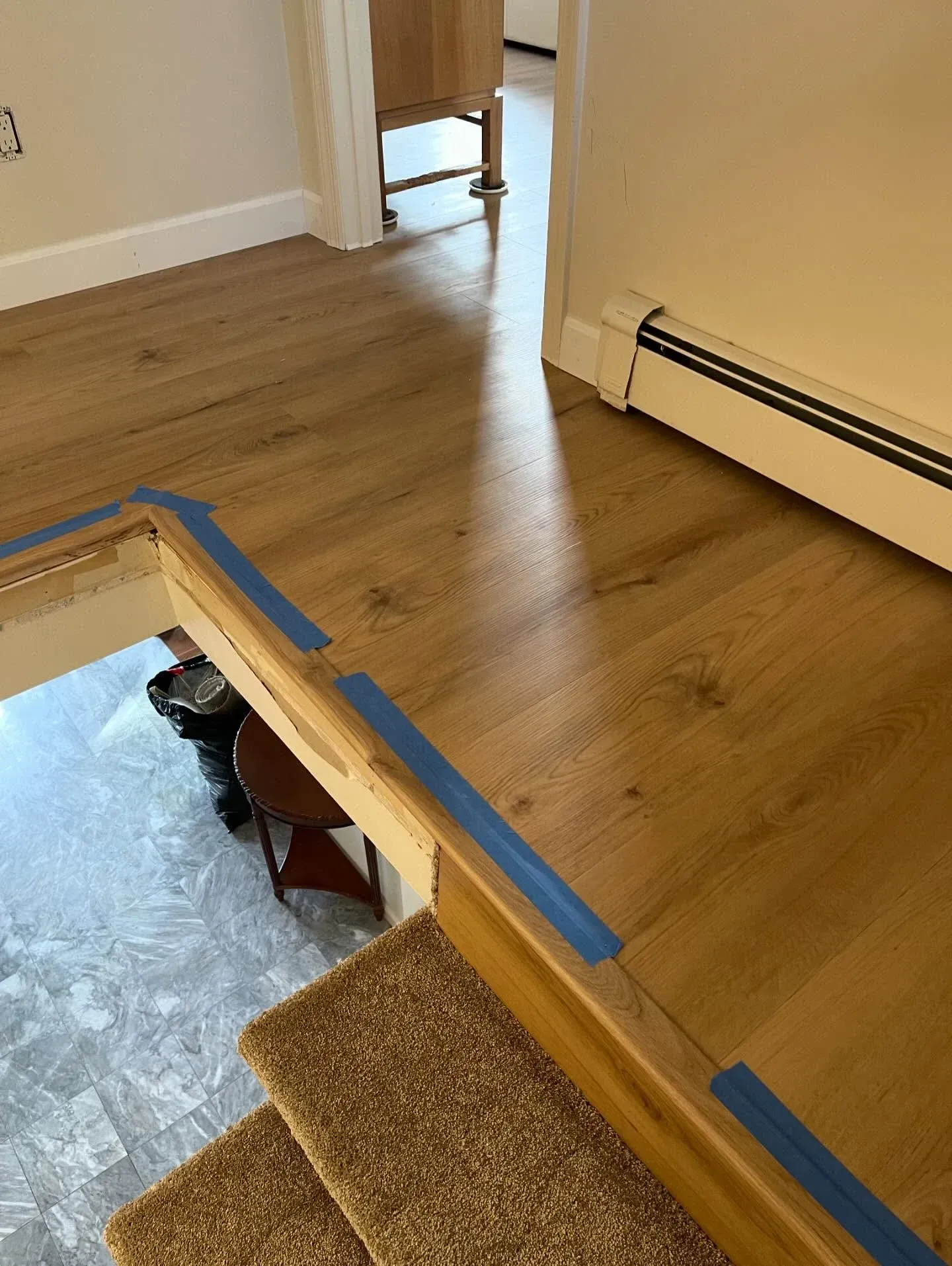 Indoor stair opening with blue tape, wooden floor, and beige baseboard heater near carpeted steps
