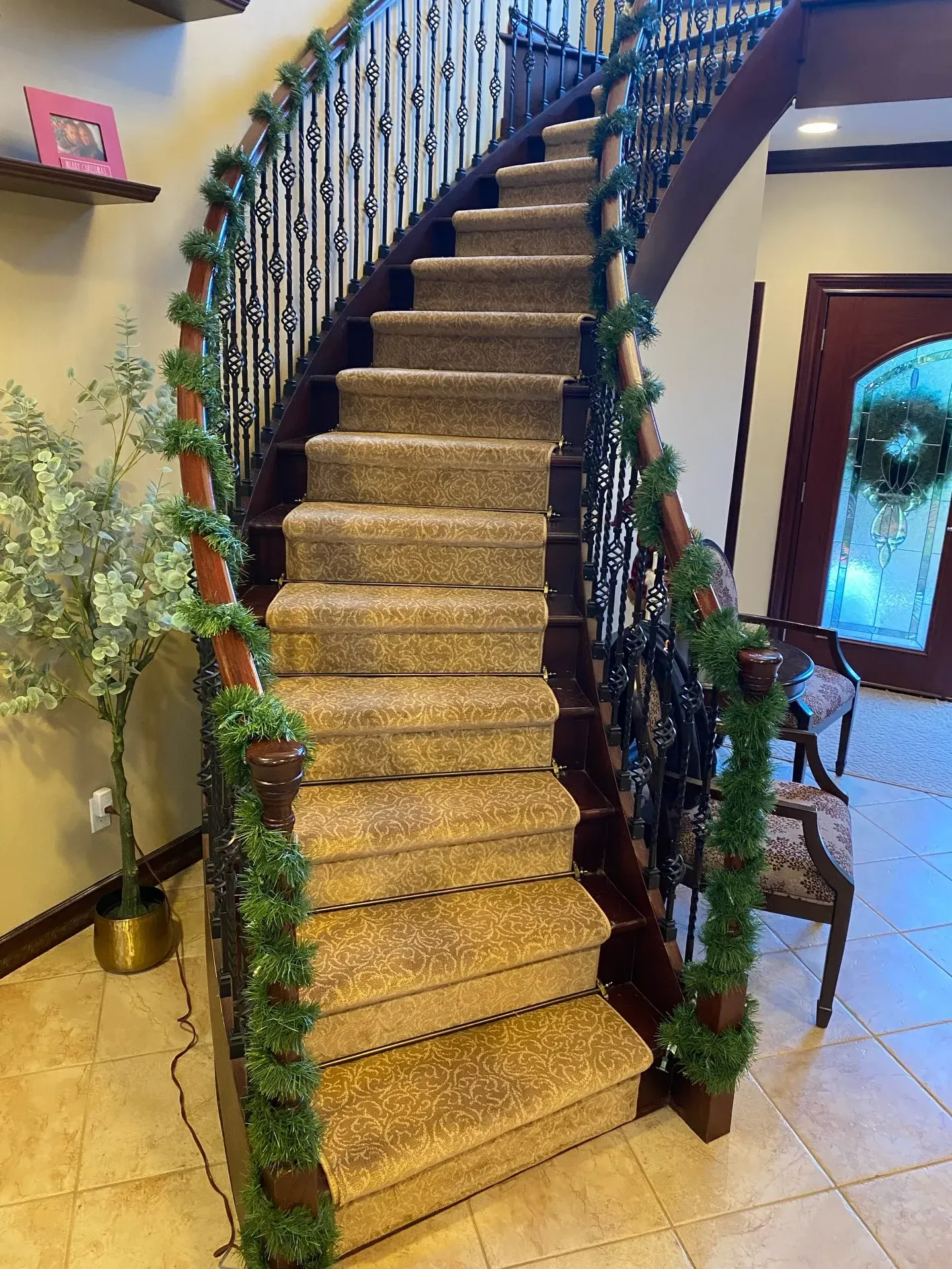 Curved carpeted staircase with black wrought-iron railing in a tiled foyer, decorated with trailing green garlands.