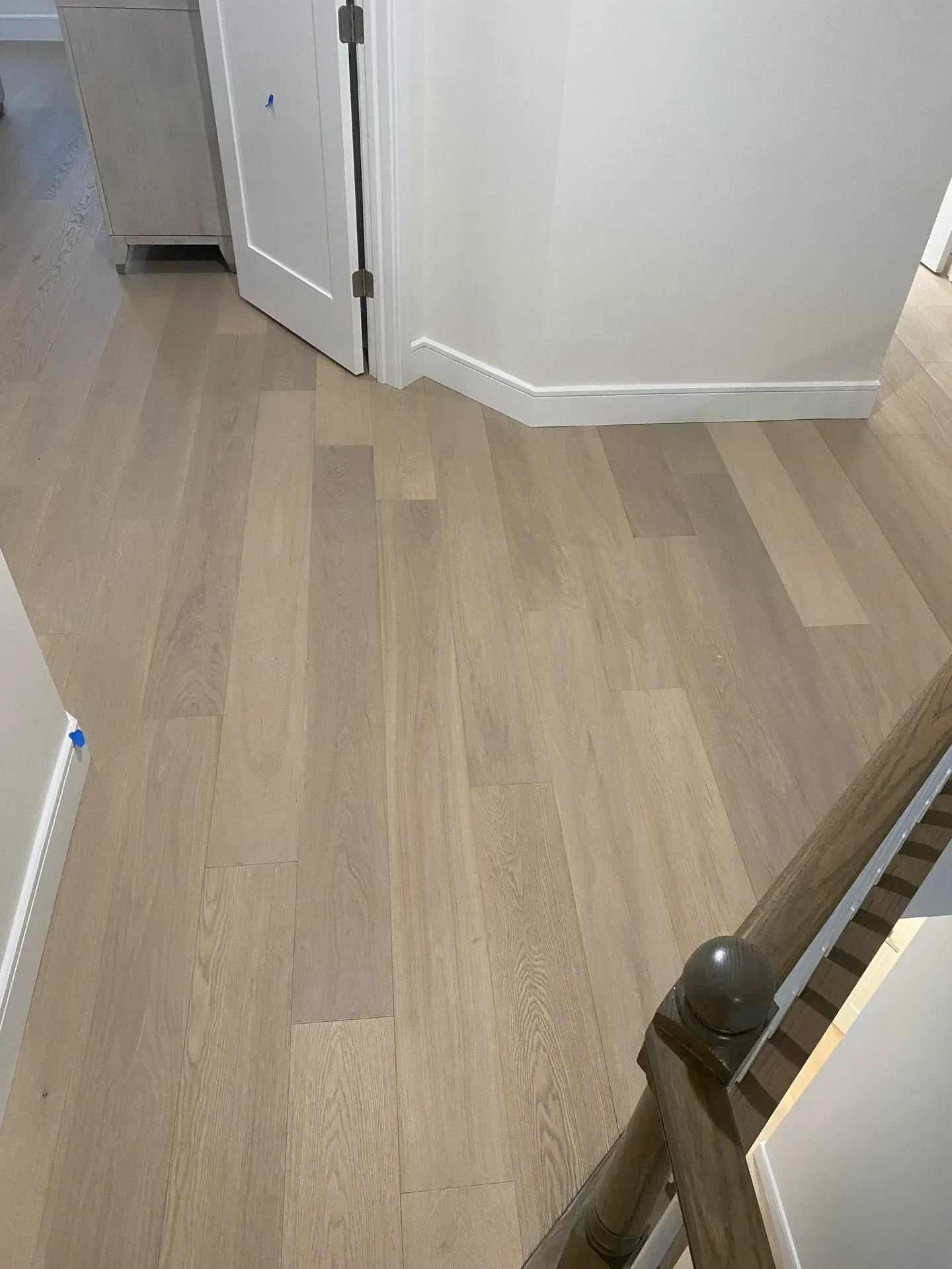 Light wood floor in a hallway with white walls and a staircase railing at right.