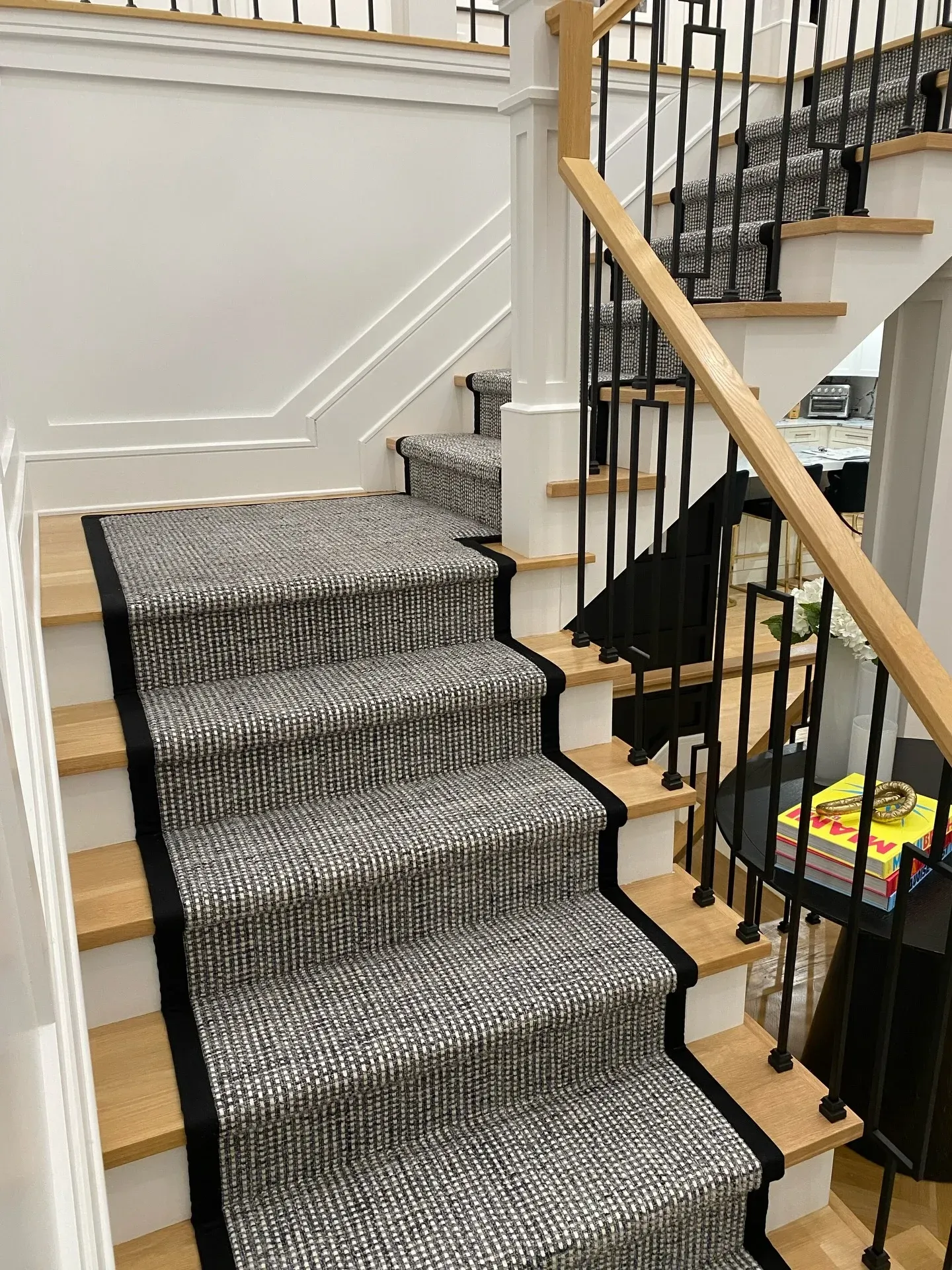 Staircase with patterned carpet runner, white walls, wood banister, and black iron railing.