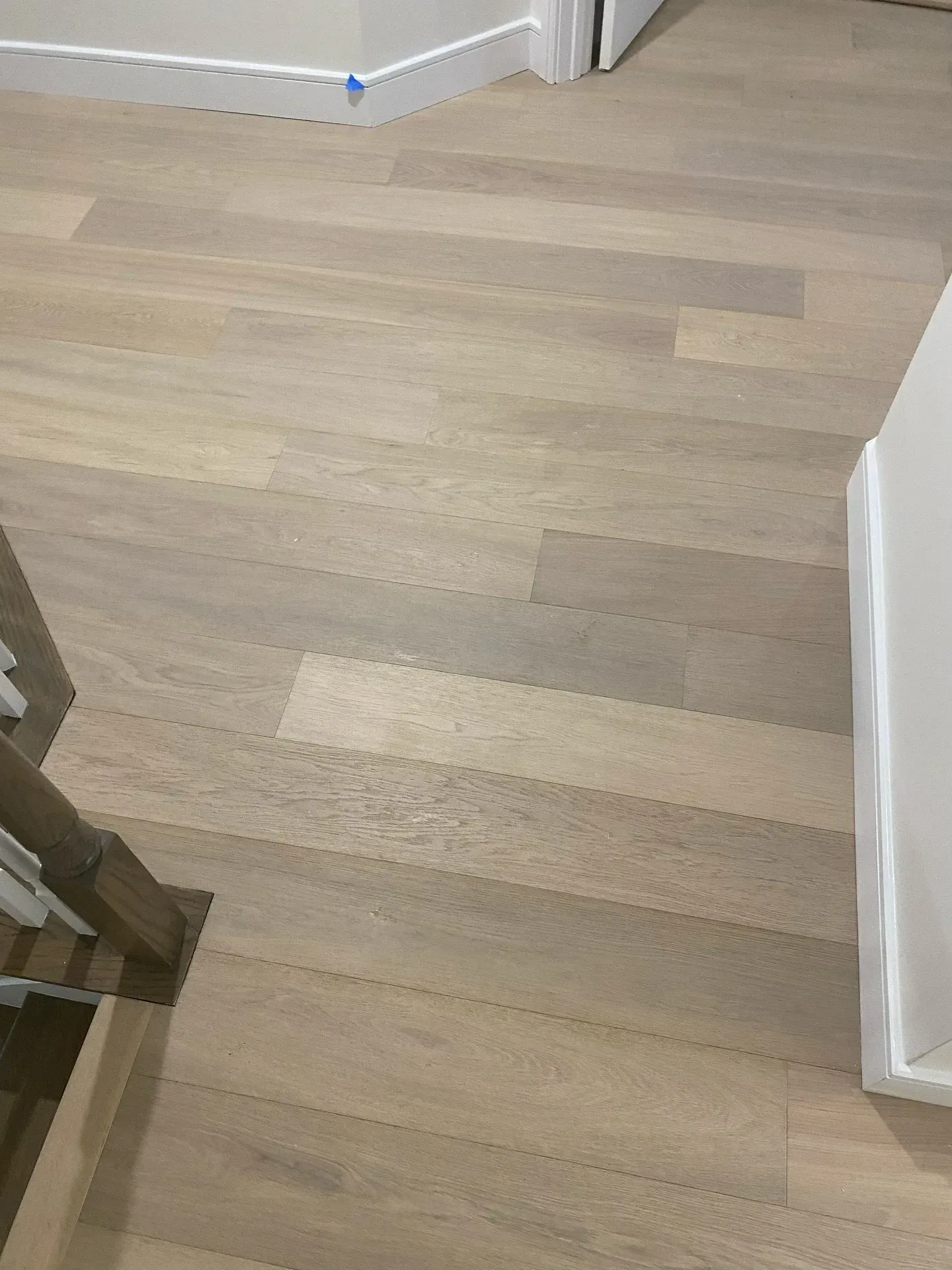 Light wood flooring in a hallway corner with white walls and a staircase railing visible.