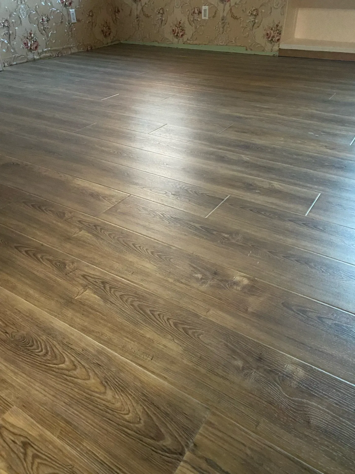 Empty room with gray-brown wood flooring and floral wallpaper, lit by a bright reflection on the floor