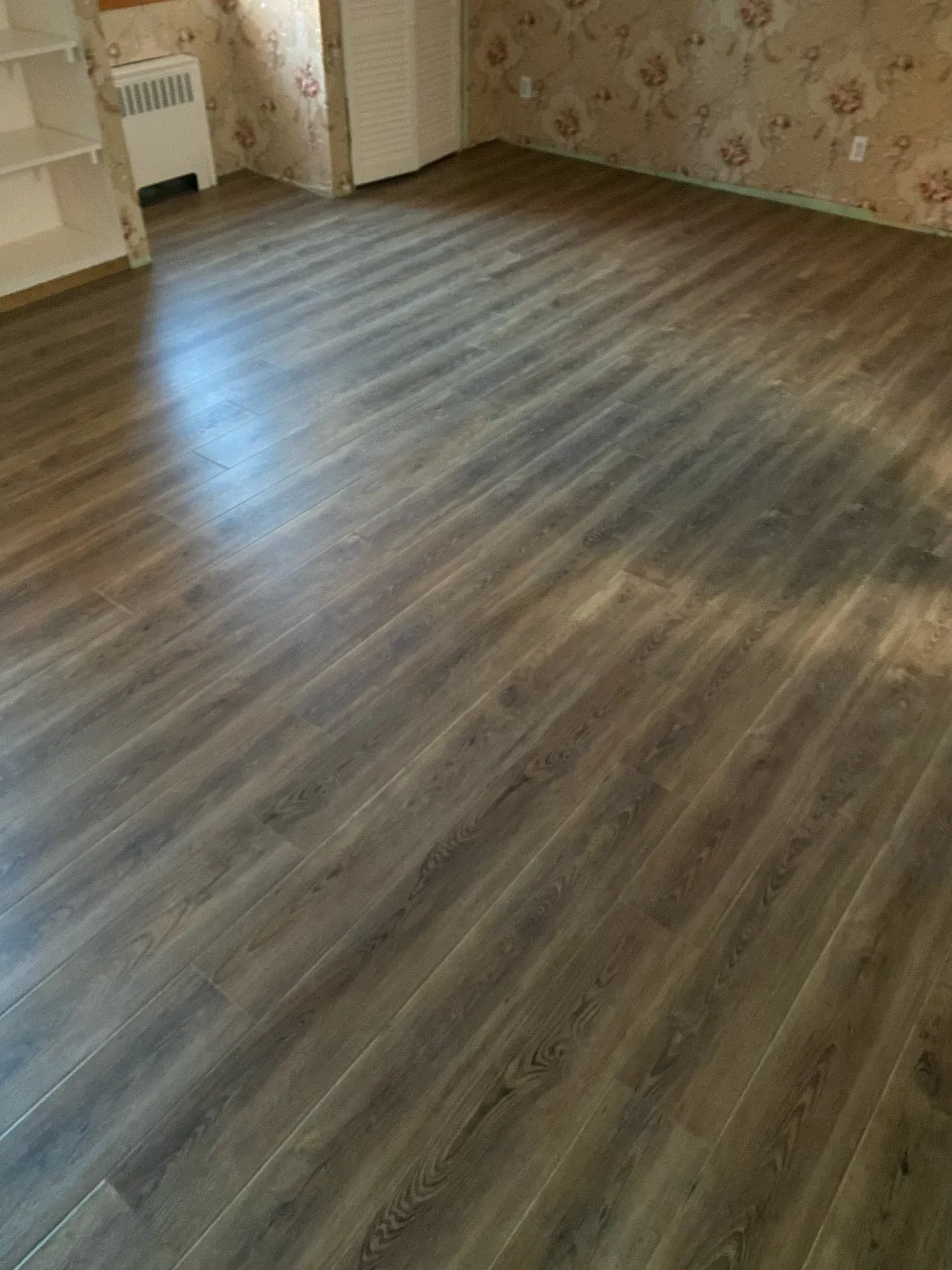 Empty room with wood-look flooring, patterned wallpaper, and a radiator near the corner