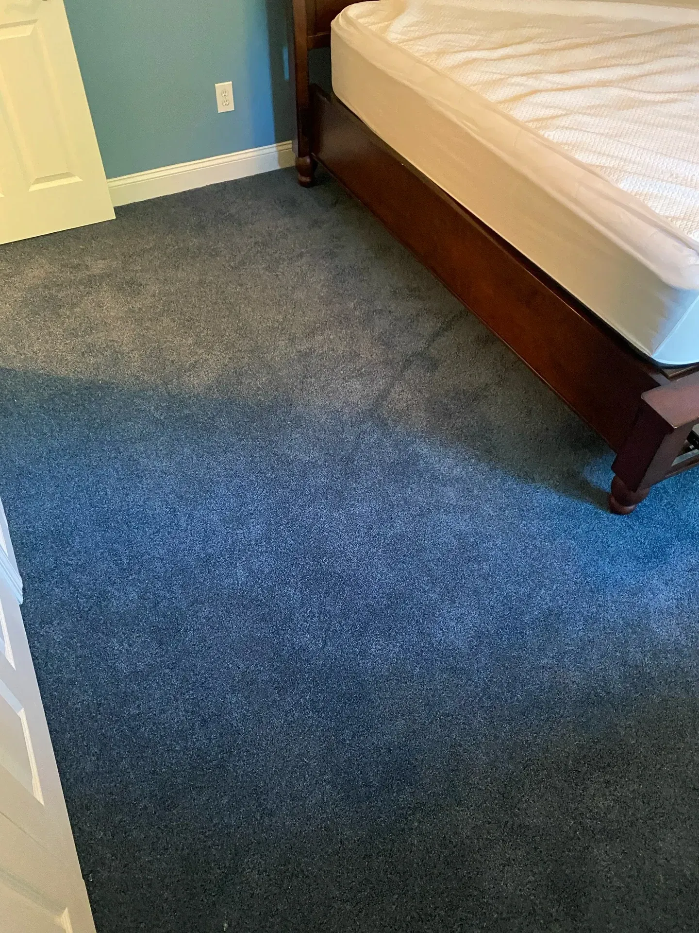 Bedroom corner with blue carpet, white door, and a wooden bed frame with a beige mattress.