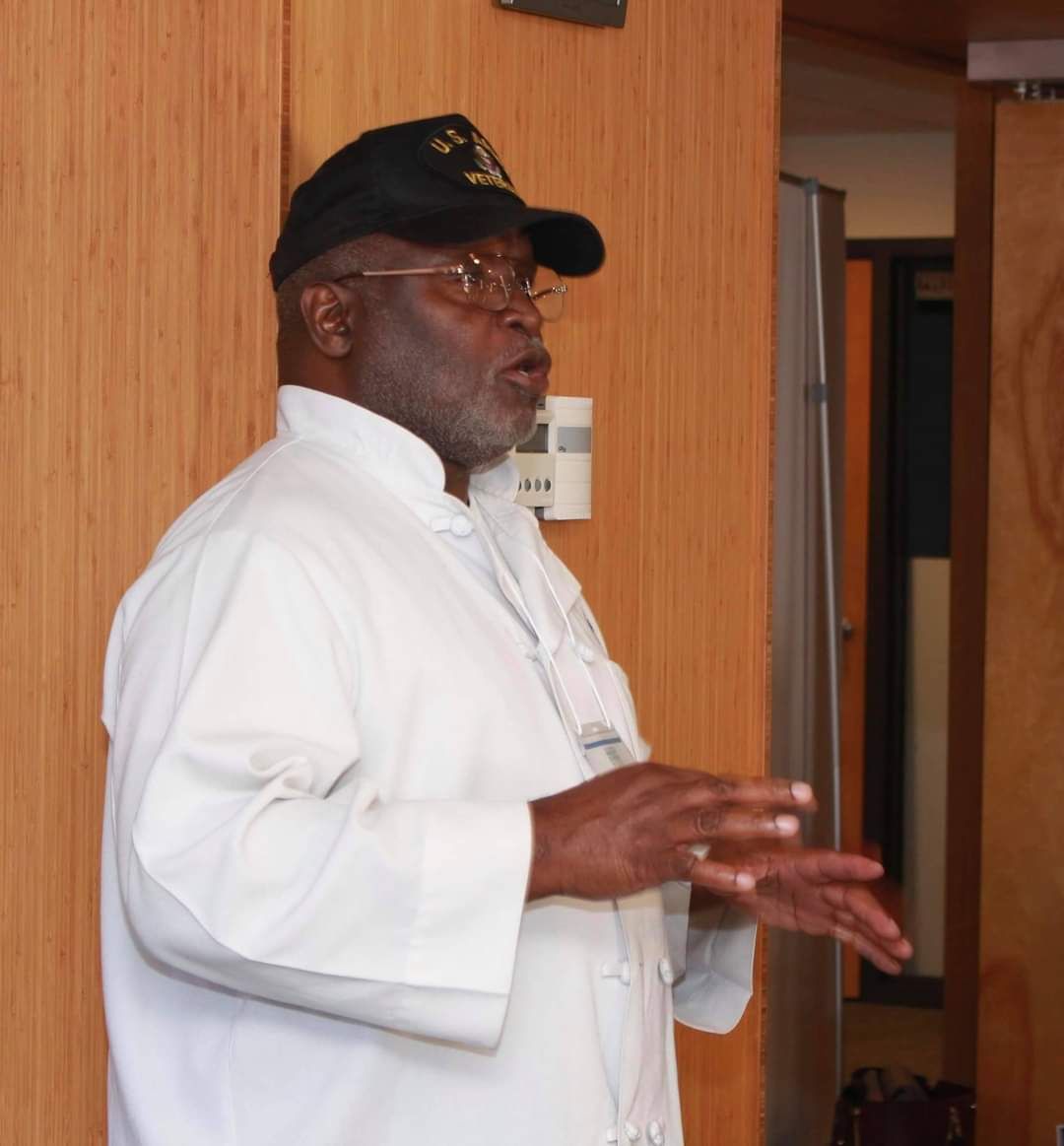 A man wearing a black hat and glasses stands in front of a wooden door