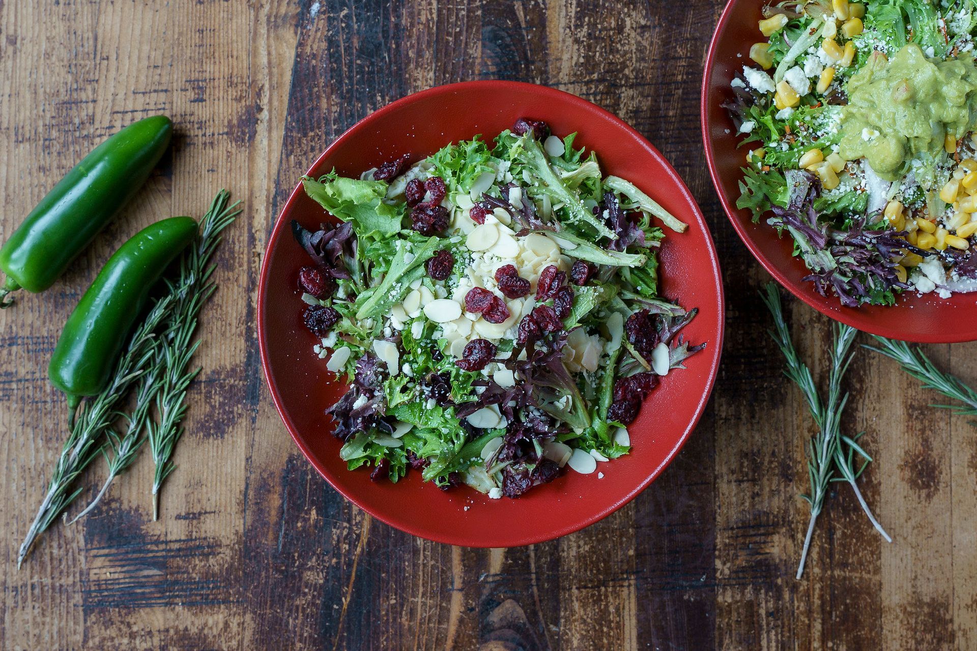Salads in red bowls, topped with cranberries and almonds, on a wooden table with jalapeños and rosemary.