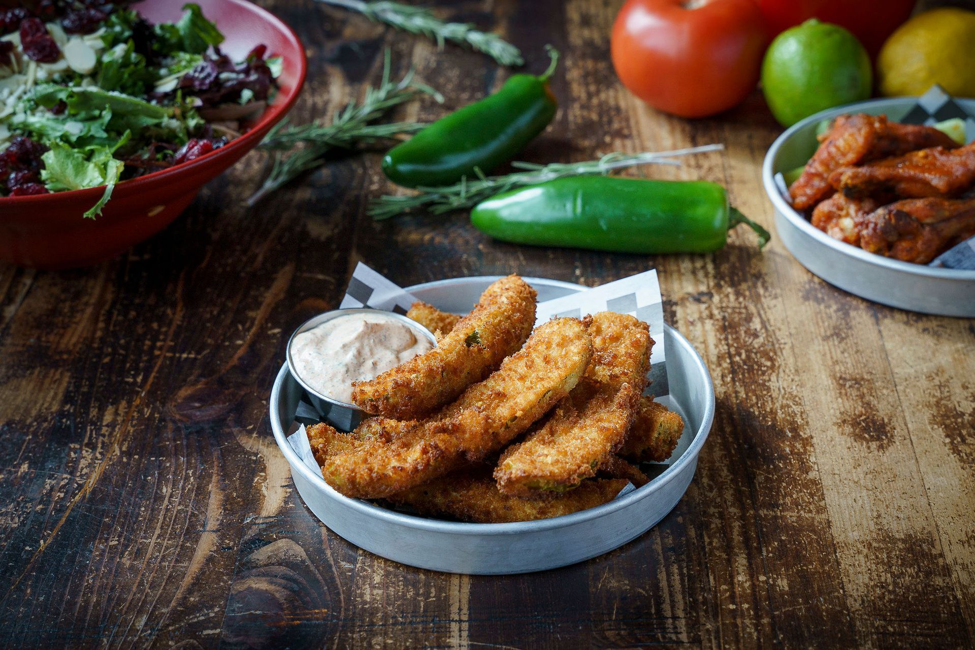 Fried pickles with dipping sauce, salad, chicken wings, and ingredients on a wooden table.