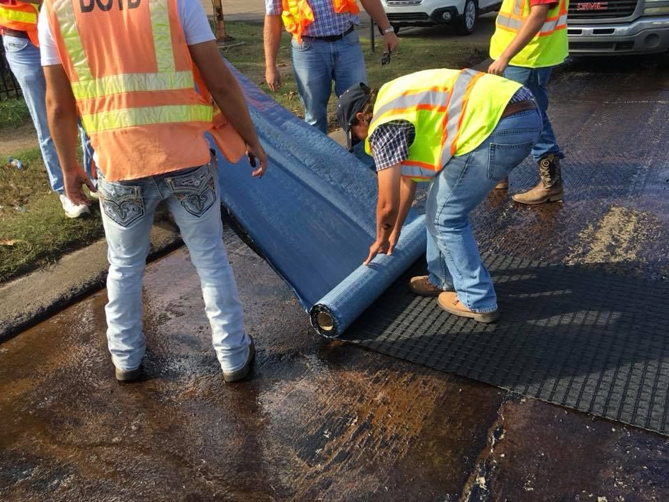 Workers rolling out blue roofing underlayment on a roof, wearing orange safety vests.