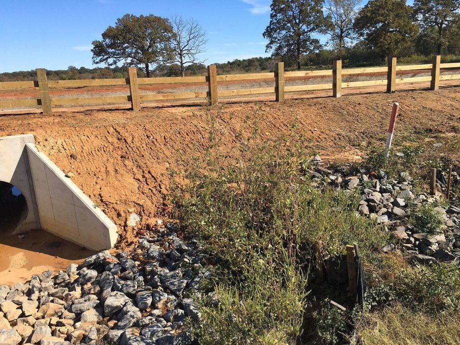 Dirt embankment with rocky drainage culvert beside a wooden fence in a rural field