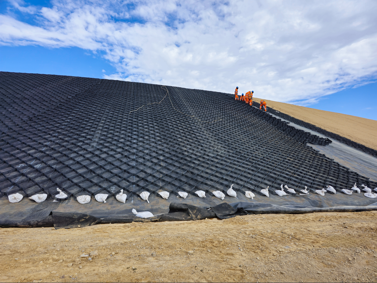 Black geomembrane-lined slope at a landfill with workers near the top under a blue sky
