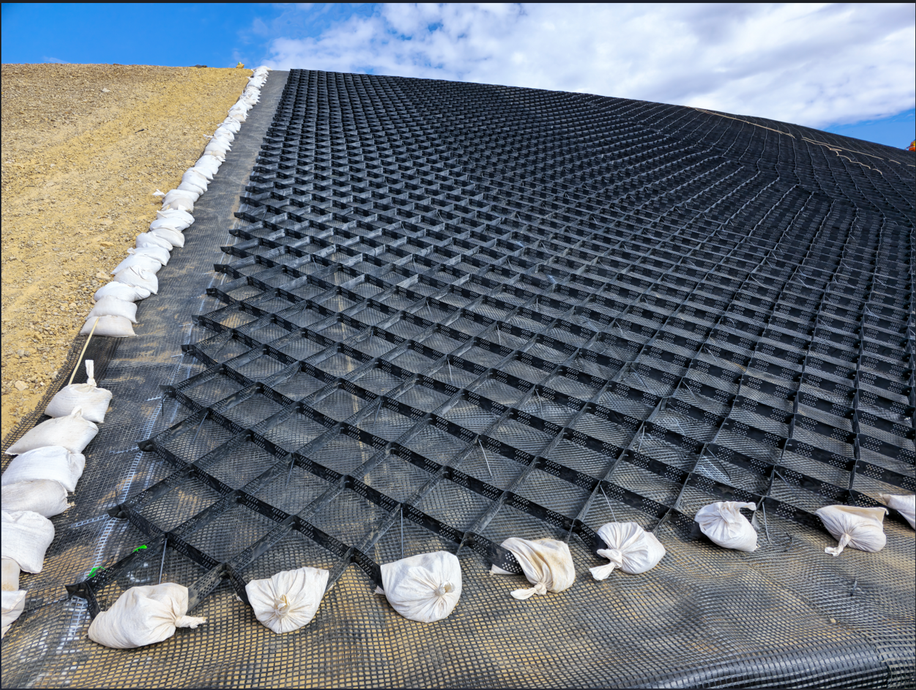 Black erosion-control mesh on a sloped dirt embankment, secured with white sandbags under a blue sky