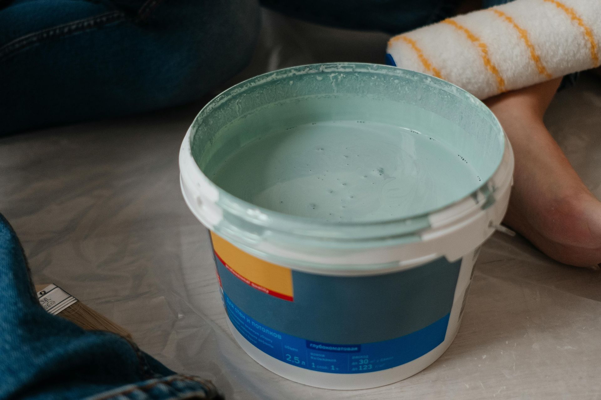 Open paint bucket with light blue paint on a floor рядом a striped sock and person’s leg