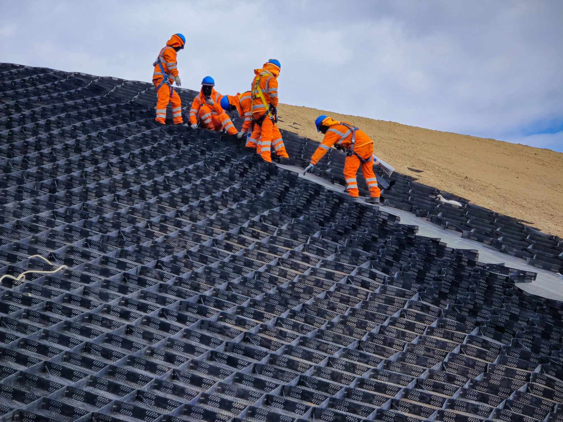 Workers in orange gear installing black geotextile netting on a sloped hillside.