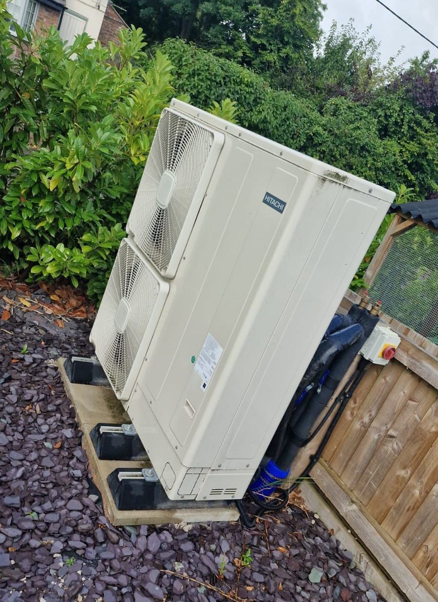 A large white air source heat pump is sitting on top of a pile of gravel next to a wooden fence.