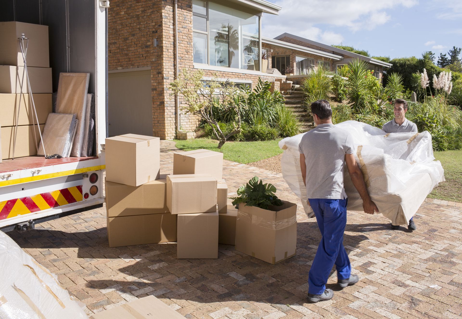 Movers carrying a wrapped sofa out of a truck, boxes scattered on a brick driveway in front of a house.