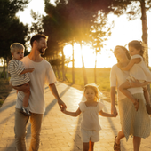 Family walking hand in hand on a sunlit path at sunset, with two children being carried.