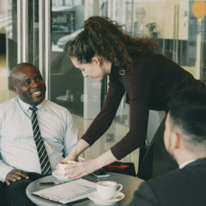 Business meeting in a glass-walled office, with a woman handing papers to a seated man at a table.