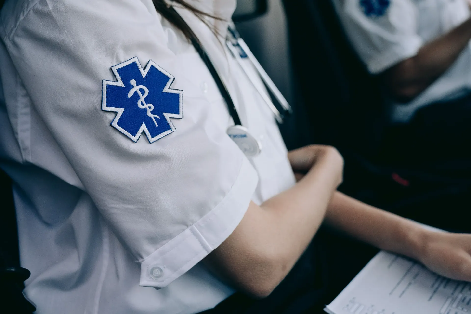 Paramedic in light blue uniform with Star of Life patch seated beside another person in a medical setting