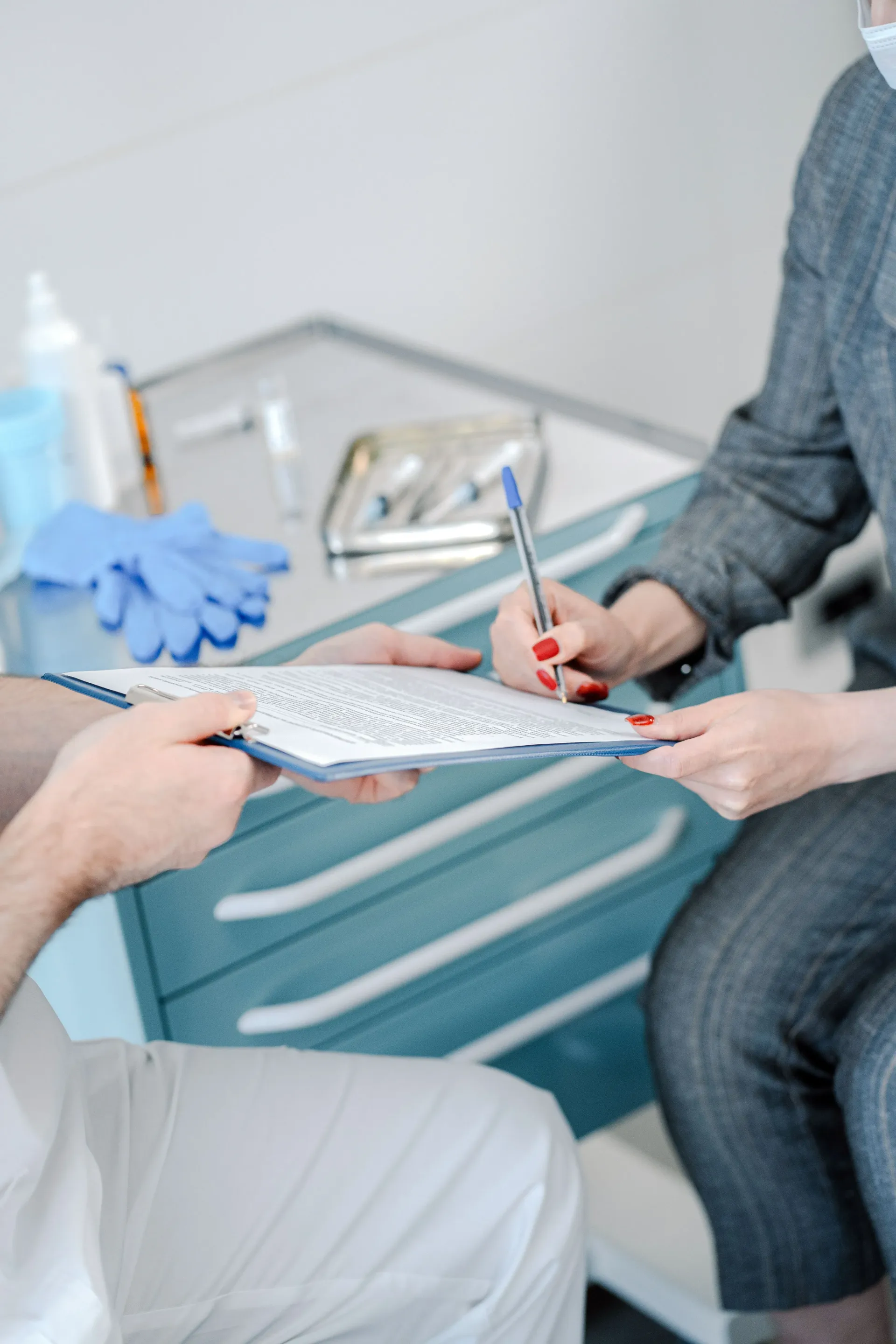Two people discuss paperwork in a clinic, one holding a pen over a clipboard near medical gloves.