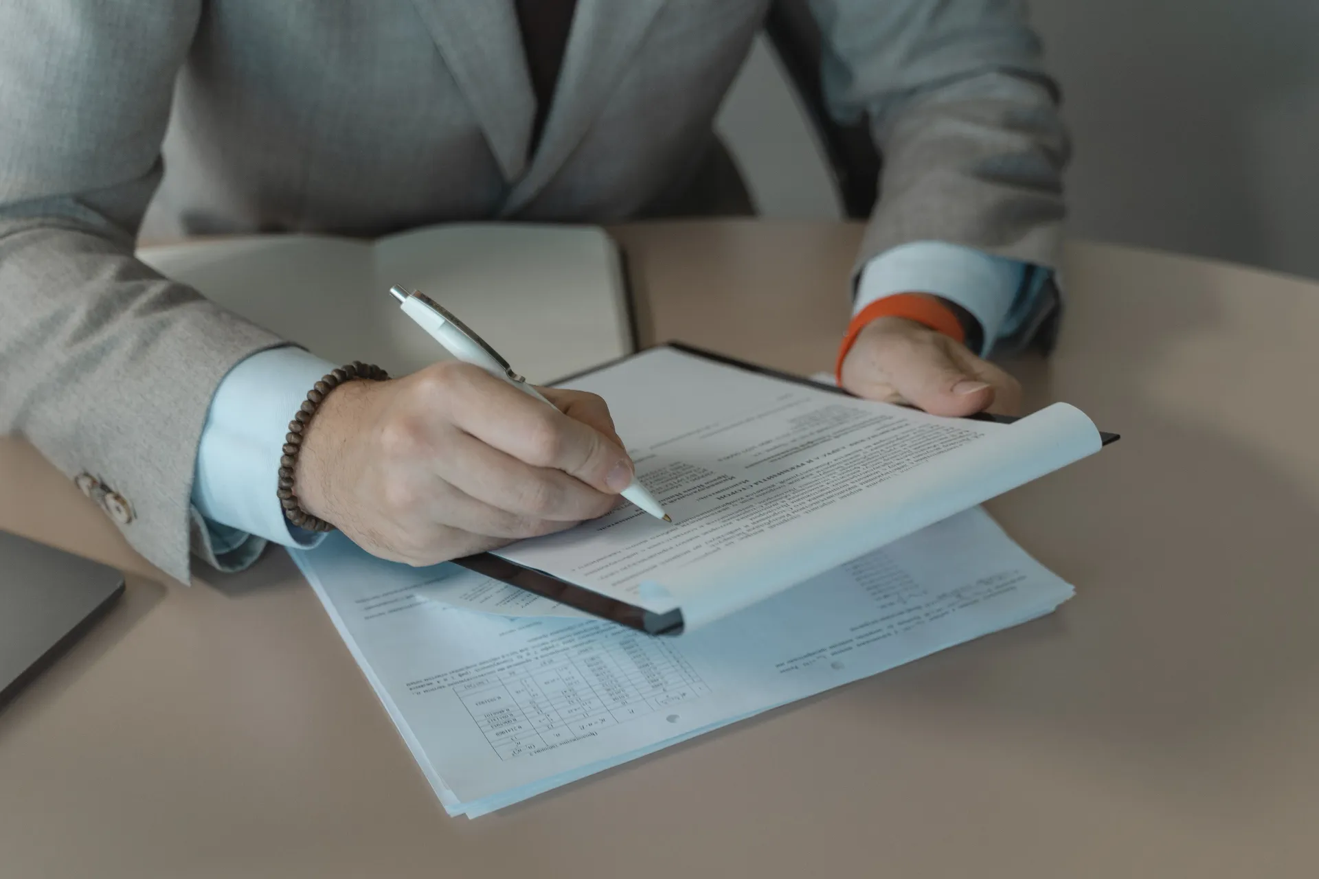 Person in a gray suit writing on a document at a desk with papers and a pen