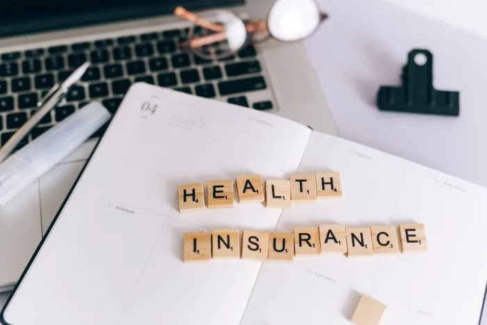 Scrabble tiles spelling “HEALTH INSURANCE” on papers beside a laptop and desk items