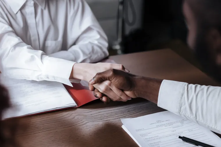 Two people shaking hands across a wooden table with papers and a pen nearby