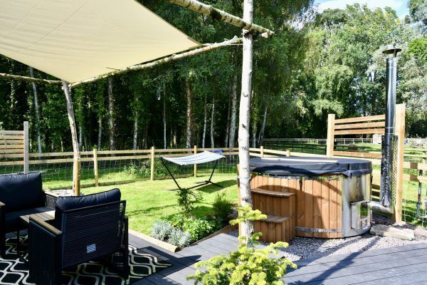Backyard deck with hot tub, patio seating, shade sail, and green trees beyond a wooden fence