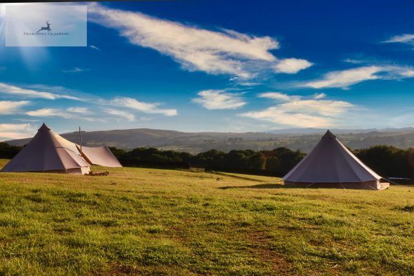 Two white tents in a grassy field under a blue sky with clouds and distant hills