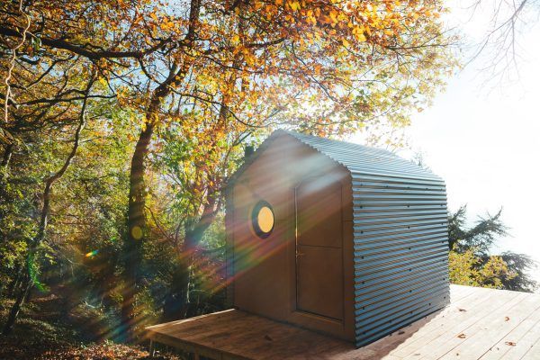 Small modern wooden cabin with a circular window, surrounded by autumn trees and sunlight