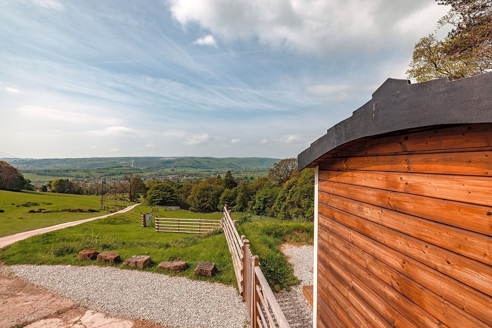 Wooden cabin beside a gravel path overlooking green hills and a wide valley under a partly cloudy sky