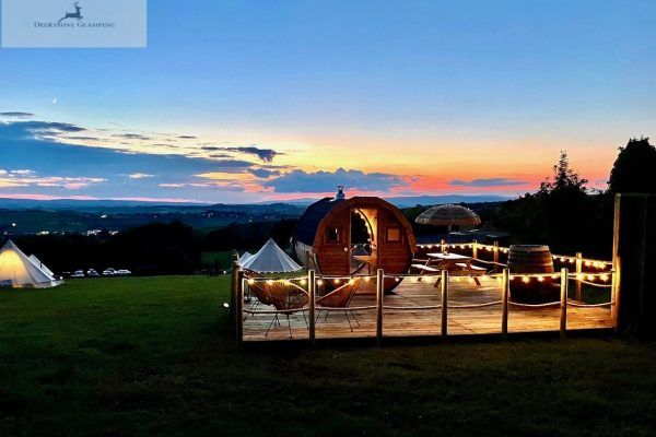 Outdoor sunset view of a lit glamping tent and deck overlooking a grassy field and distant hills