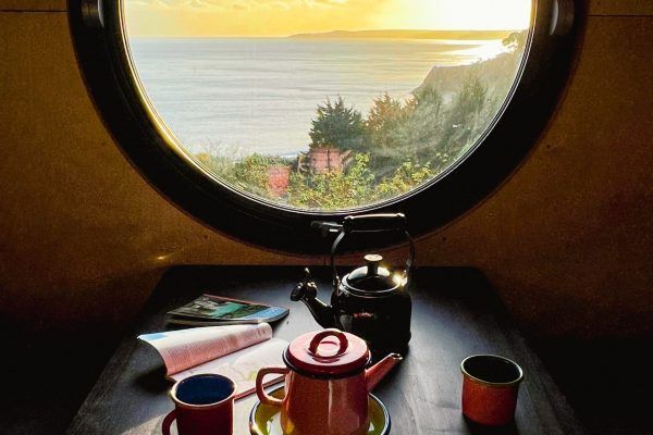 Sunlit ocean view through round window above a table with teapot, cups, and books