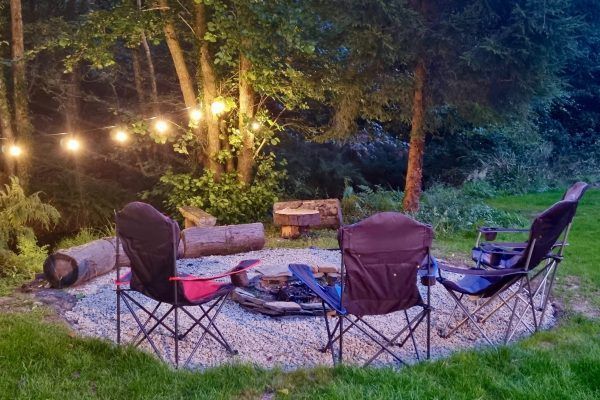 Campfire seating area in a wooded backyard at dusk, with string lights, chairs, and logs around a fire pit.