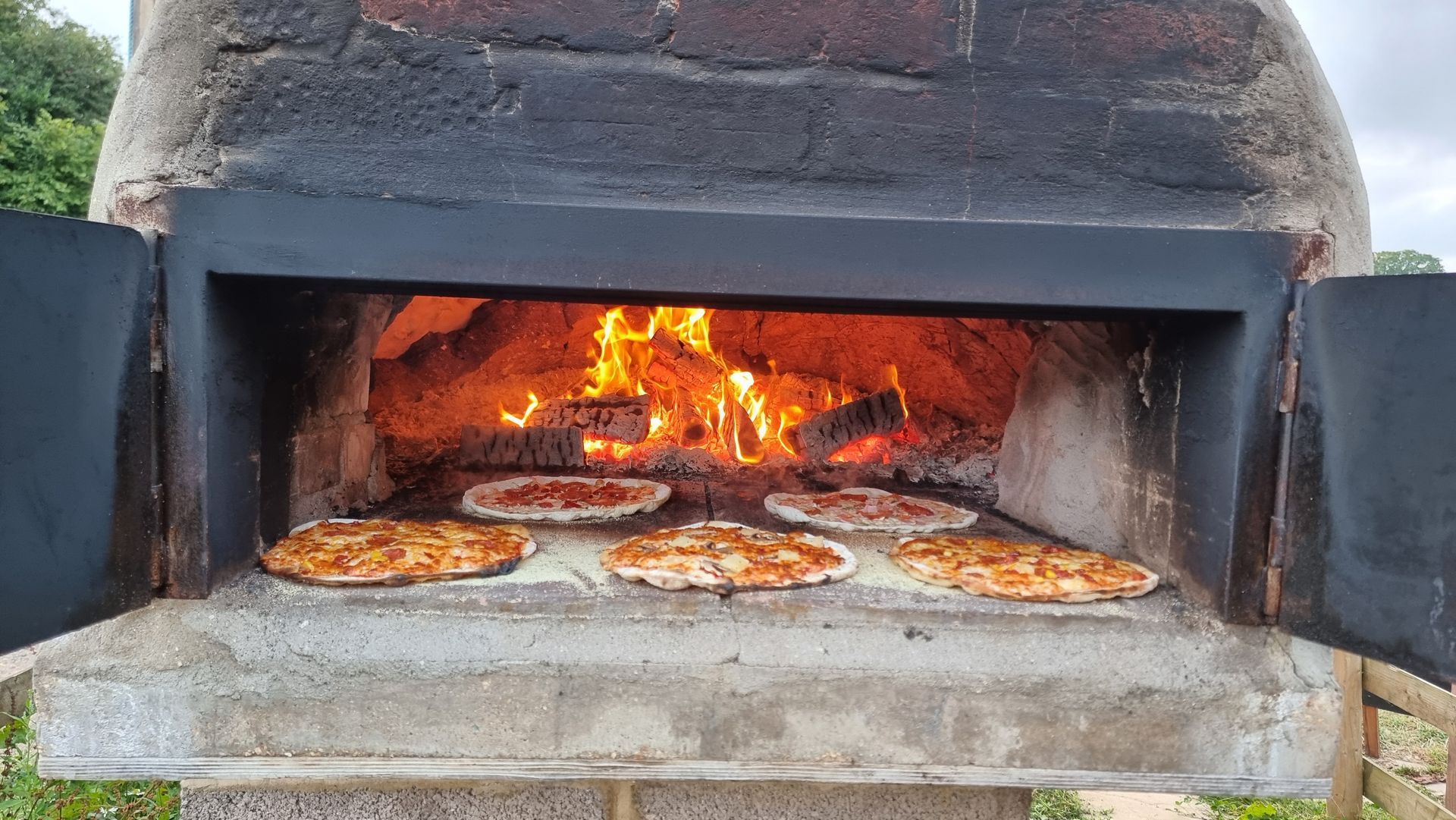 Wood-fired oven with pizzas baking inside, flames visible in the back