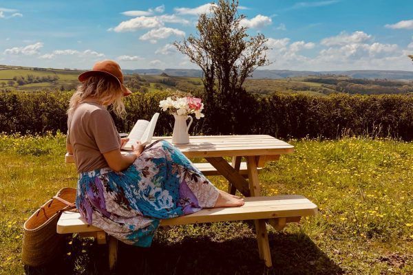 Person sitting at a picnic table in a field, writing with flowers and a scenic countryside view beyond.