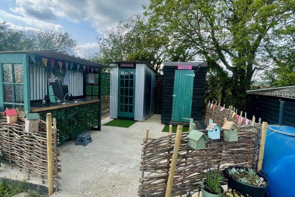 Small garden shed and patio area with woven fence, potted plants, and trees overhead