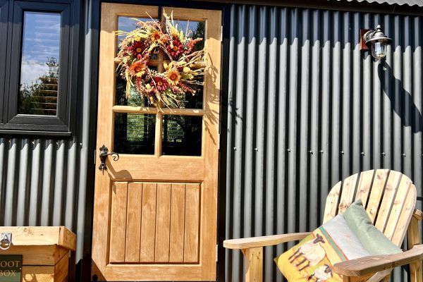 Wooden door with autumn wreath on corrugated metal wall beside a wicker chair with cushion.