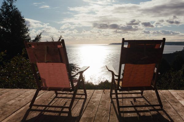 Two empty deck chairs facing a bright lake at sunset, with trees and cloudy sky.