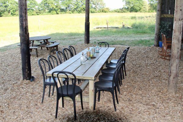 Outdoor picnic area with a long wooden table, black chairs, and trees overlooking a grassy field.