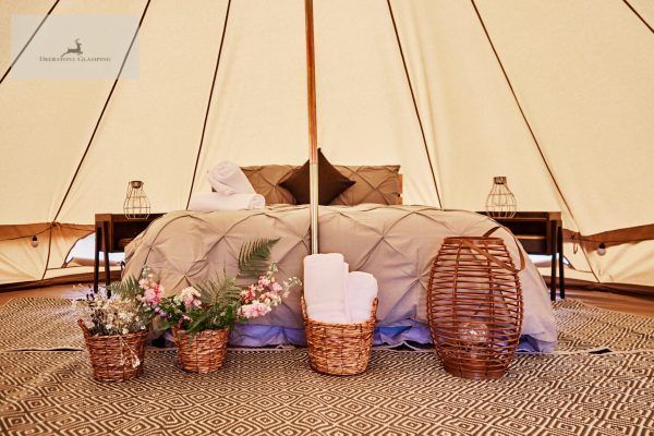 Cozy bed inside a beige canvas tent, with pillows, blankets, and woven baskets and flowers in front