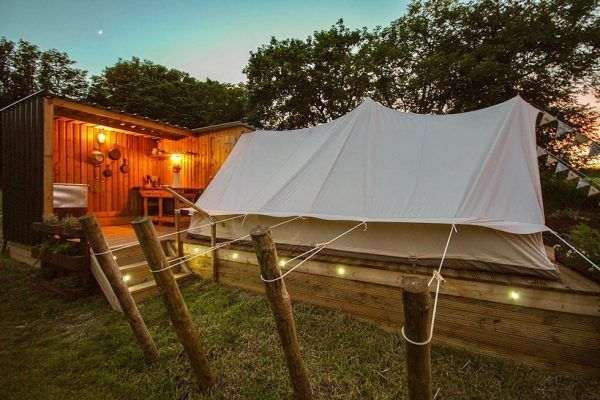 Wooden cabin beside a white canvas tent at dusk, with rope fence and trees in the background