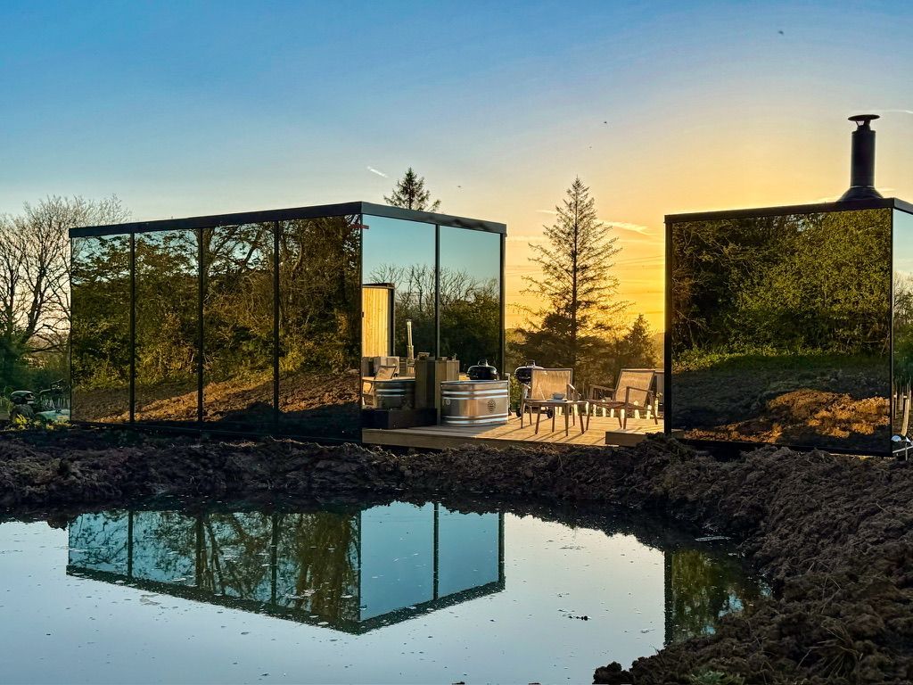 Modern glass cabin at sunset, reflected in a still pond, with outdoor seating and warm golden light