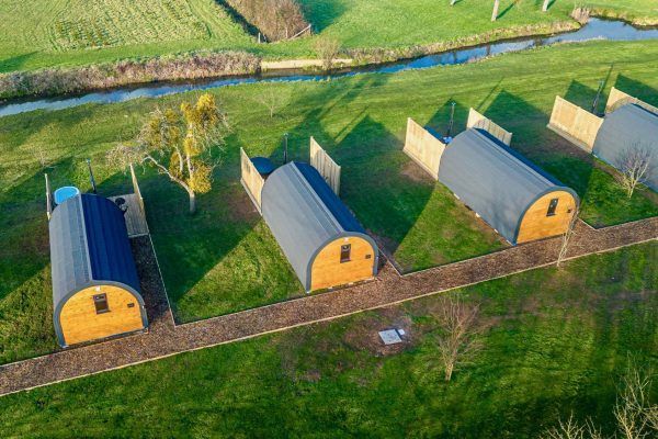 Aerial view of three small arched-roof cabins on green grass beside a road and stream.