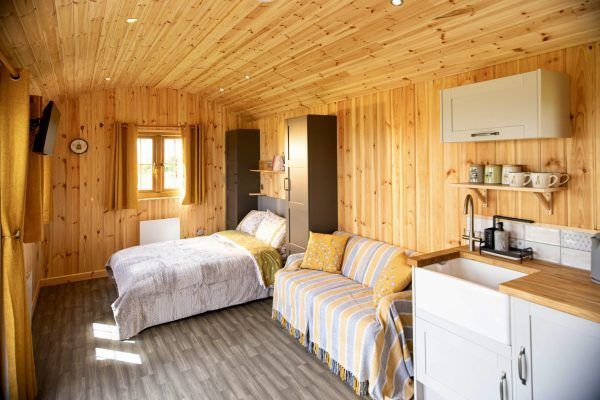 Cozy wooden cabin interior with a bed, striped sofa, small kitchen, and open doorway to a bright outside view