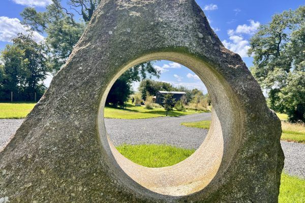 Stone sculpture with a circular opening in a sunny park, framed by green grass and trees.