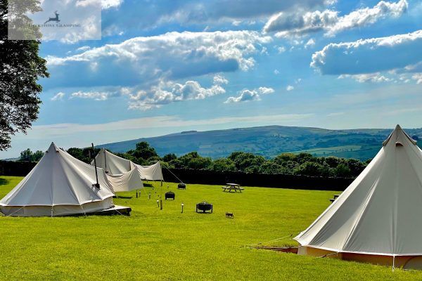 White canvas tents on a green hillside overlooking distant mountains under a partly cloudy blue sky