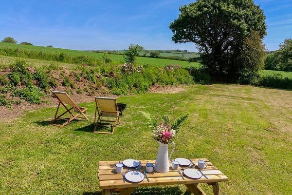 Picnic setup on a grassy field with wooden chairs, a set table, and a tree under a clear blue sky.